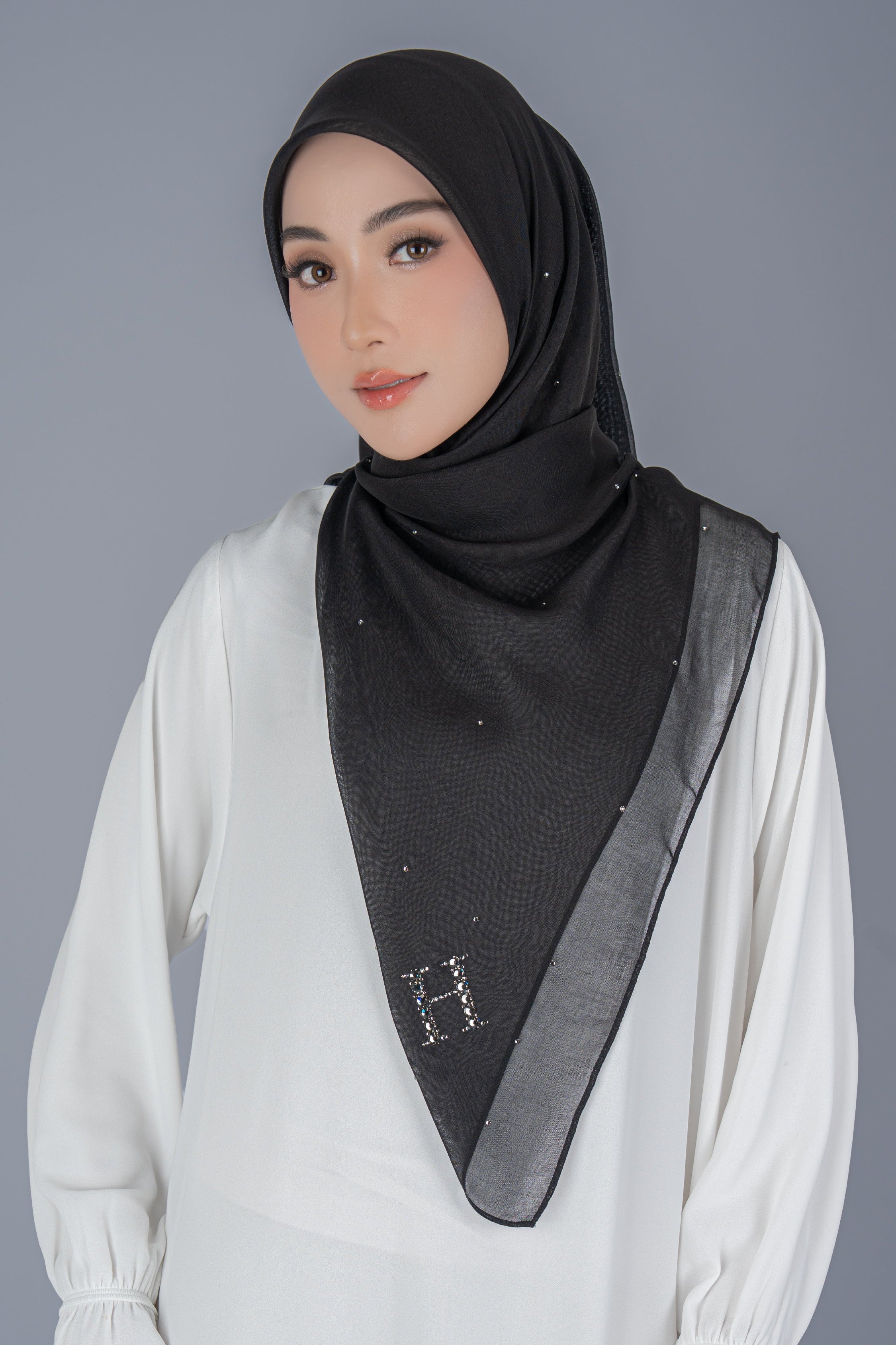 A woman wearing a black hijab with a wide gray border and small silver rhinestone accents, paired with a white long-sleeve top, against a solid gray studio background.